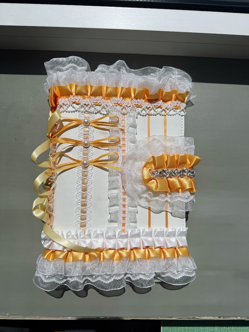 May include: A white and yellow decorative journal with lace and ribbon accents. The journal features a ruffled trim, satin ribbons, and pearl embellishments. The design includes a matching accessory with a jeweled band.