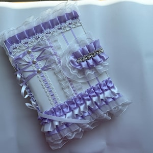 May include: A white and lavender-colored notebook cover with decorative ribbons, pearls, and lace. The cover features a ruffled edge and a small, matching accessory with a rhinestone accent. The design is ornate and feminine.