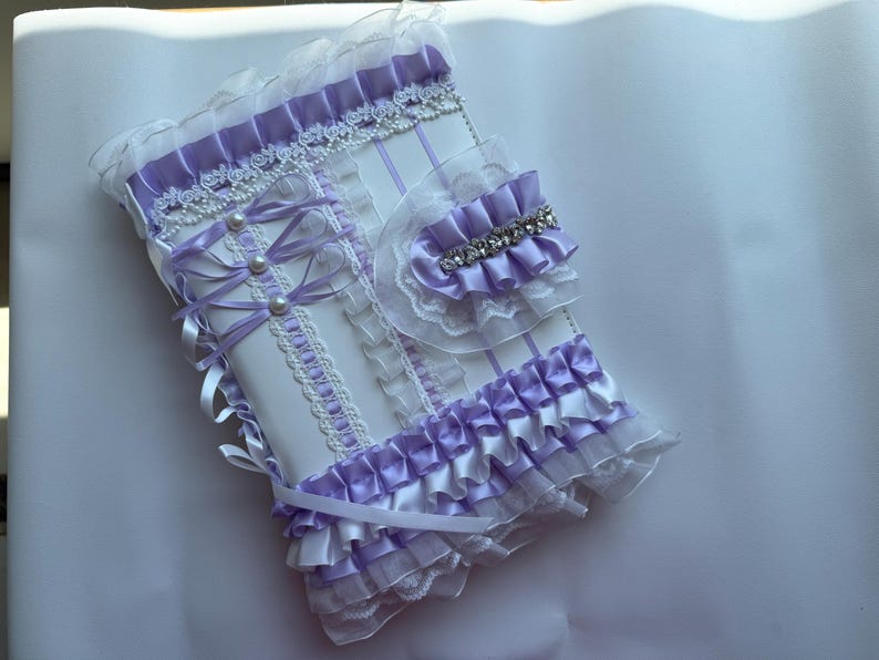 May include: A white and lavender-colored notebook cover with decorative ribbons, pearls, and lace. The cover features a ruffled edge and a small, matching accessory with a rhinestone accent. The design is ornate and feminine.
