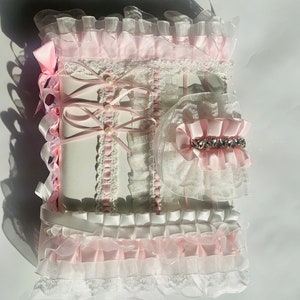 May include: A decorative, frilly, white and pink journal with a matching accessory. The journal is adorned with layers of lace, ribbons, and pearls. The accessory features a pink bow and a sparkling rhinestone embellishment.