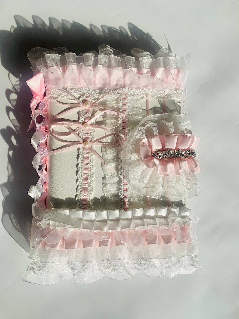 May include: A decorative, frilly, white and pink journal with a matching accessory. The journal is adorned with layers of lace, ribbons, and pearls. The accessory features a pink bow and a sparkling rhinestone embellishment.