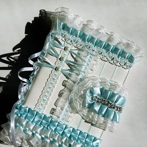 May include: A white and light blue decorative set featuring a journal and a matching accessory. The journal has a white cover with light blue ribbons, lace, and pearl accents. The accessory has a light blue bow with a rhinestone embellishment.