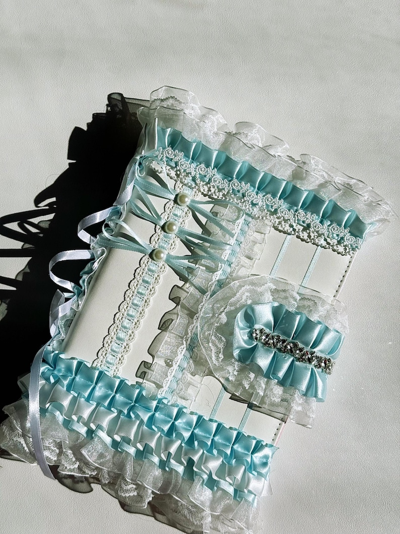 May include: A white and light blue decorative set featuring a journal and a matching accessory. The journal has a white cover with light blue ribbons, lace, and pearl accents. The accessory has a light blue bow with a rhinestone embellishment.