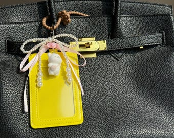 May include: Black leather handbag with a gold-tone clasp. A yellow ID holder with a clear window hangs from the handle, adorned with a white pearl bow, pink ribbon, and a small white bear charm.
