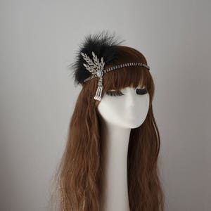 May include: A black feather headband with a silver and crystal embellishment. The headband has a beaded chain and a dangling pearl tassel.
