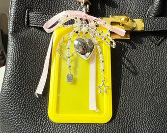 May include: A yellow phone case with a pink ribbon and a silver heart charm, pearl beads, and a star charm. The case is attached to a black leather handbag with a gold clasp.