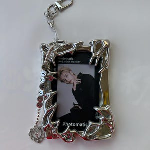 May include: Silver photo frame keychain with a photo of a man inside. The frame has a decorative design and a chain with a heart charm. The text "Photomatic Take Your Memkry" is printed on the frame.