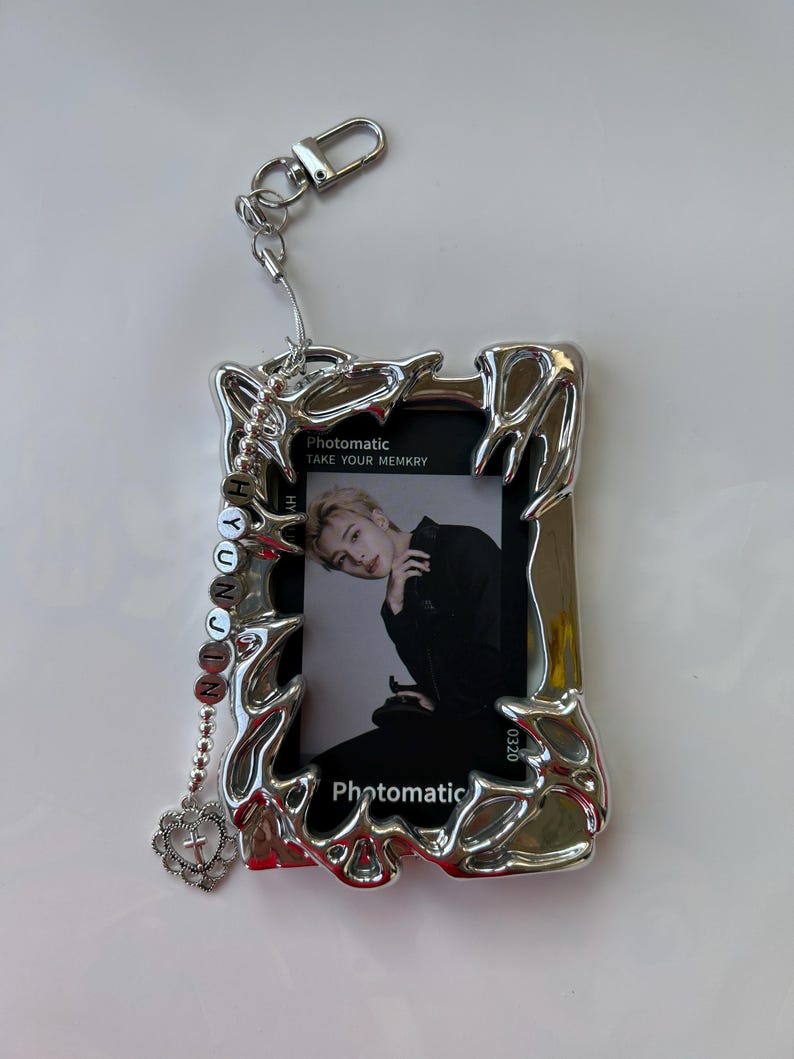 May include: Silver photo frame keychain with a photo of a man inside. The frame has a decorative design and a chain with a heart charm. The text "Photomatic Take Your Memkry" is printed on the frame.