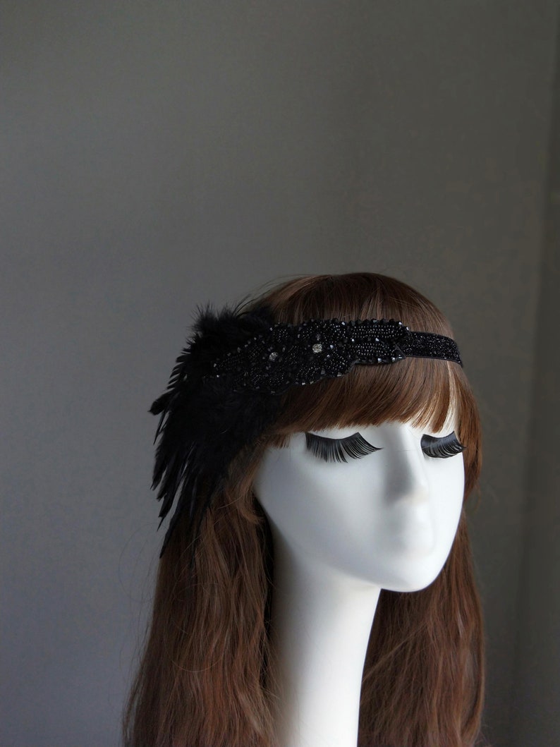 May include: A black headband with a feather and beaded embellishments. The headband is worn on a mannequin with long brown hair.