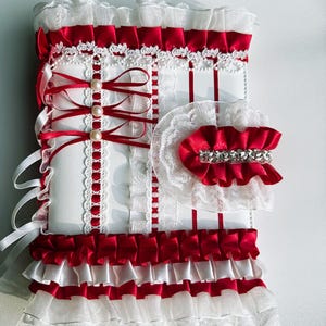 May include: A decorative white and red accessory with layers of ruffles, ribbons, and lace. The design features red satin ribbons, white lace trim, and faux pearl accents. The accessory has a bow and a rhinestone embellishment.
