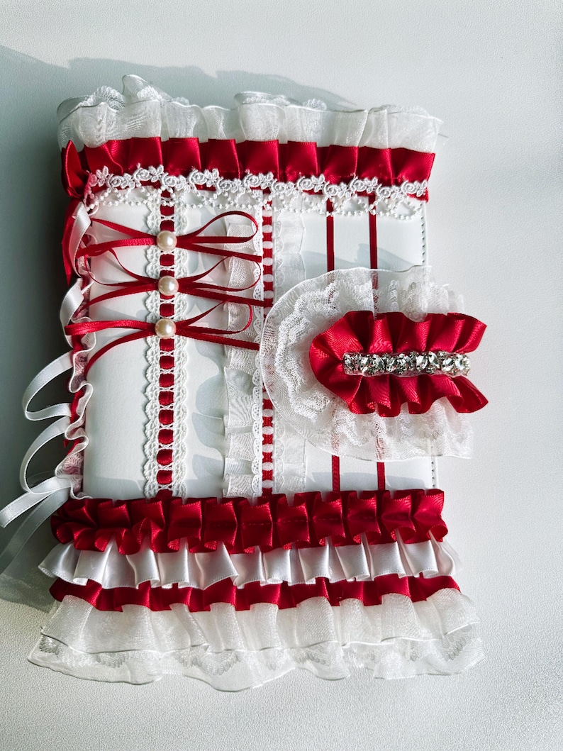 May include: A decorative white and red accessory with layers of ruffles, ribbons, and lace. The design features red satin ribbons, white lace trim, and faux pearl accents. The accessory has a bow and a rhinestone embellishment.