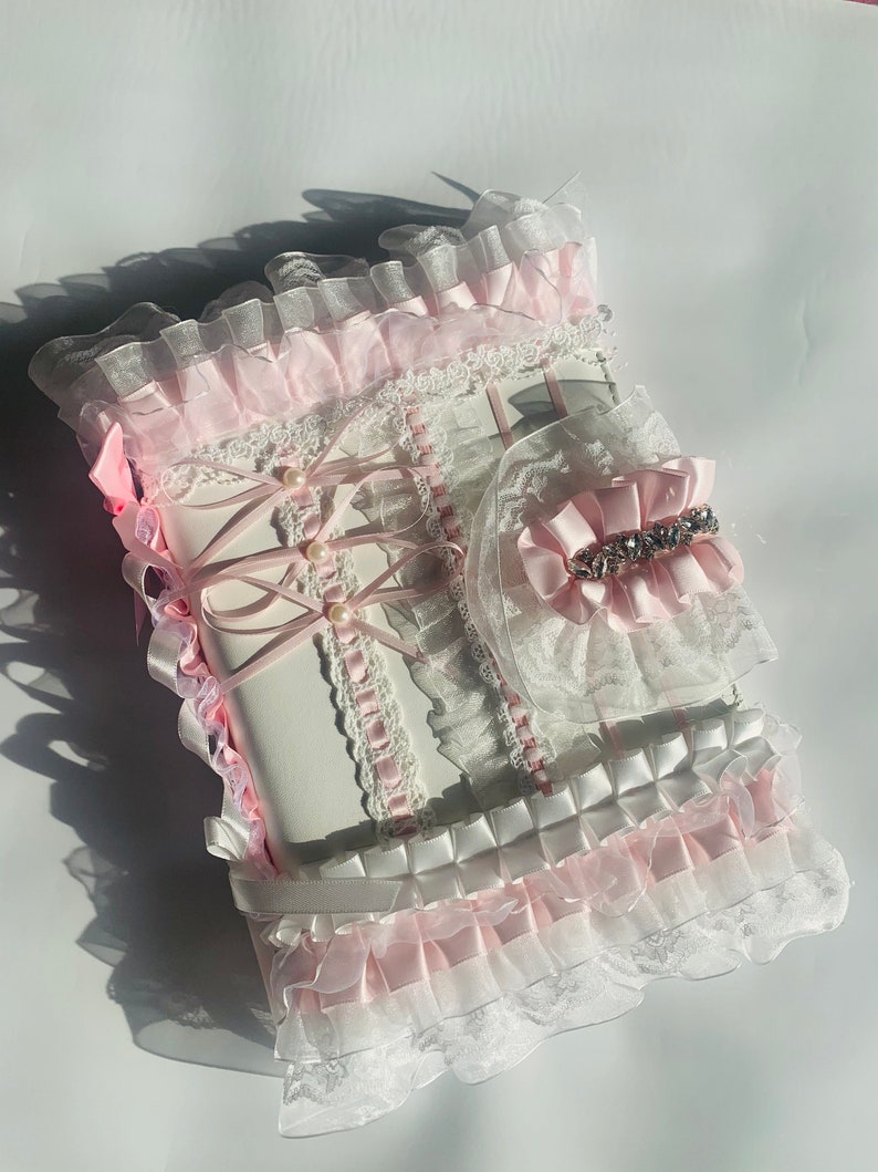 May include: A white and pink decorative journal with lace, ribbons, and pearl accents. The journal features a ruffled edge and a matching accessory with a rhinestone embellishment. The design is ornate and feminine.