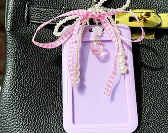 May include: A light purple ID holder with a pink and white gingham ribbon bow, pearl strands, and a decorative pink charm. The holder is attached to a black leather bag with a gold clasp.