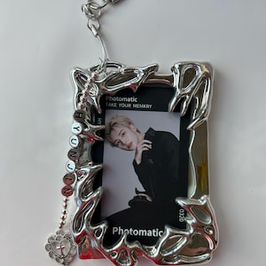 May include: Silver photo frame keychain with a photo card inside. The photo card features a young man with dark hair and a black shirt. The frame has a decorative design and a small charm with a cross hanging from it. The photo card has the text "Photomatic Take Your Memkry" and "0320".