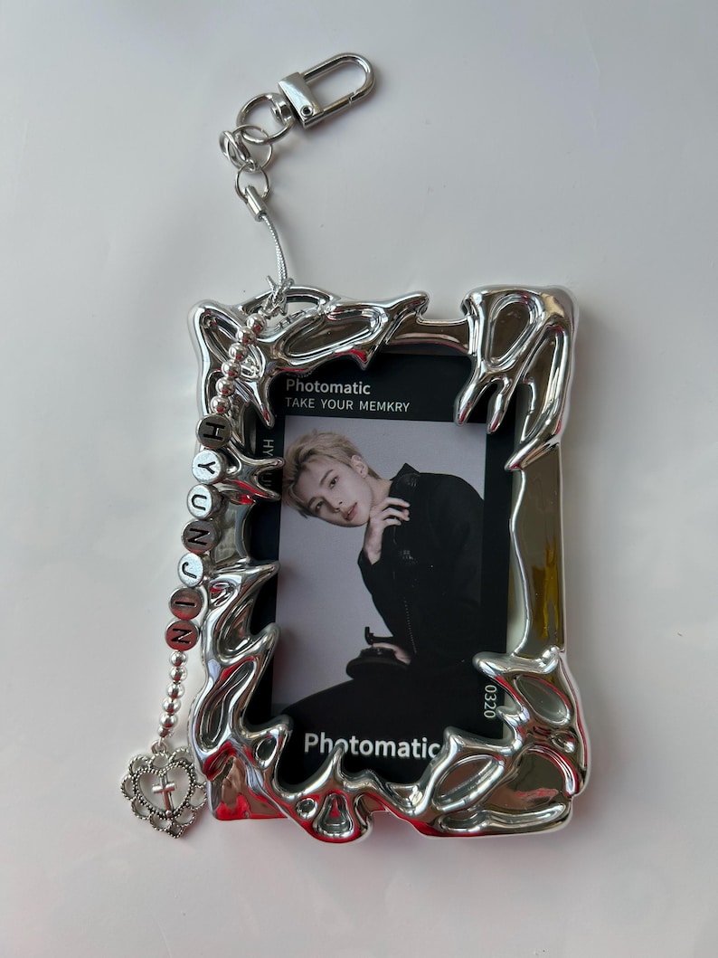 May include: Silver photo frame keychain with a photo card inside. The photo card features a young man with dark hair and a black shirt. The frame has a decorative design and a small charm with a cross hanging from it. The photo card has the text "Photomatic Take Your Memkry" and "0320".
