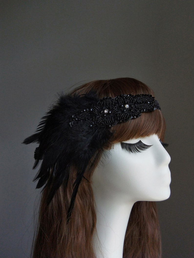 May include: A black headband with a feather and beaded embellishments. The headband is worn on a mannequin with long brown hair.