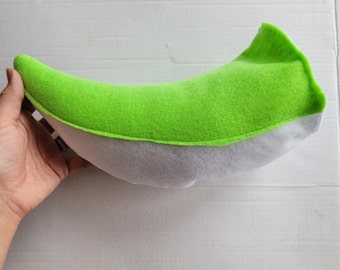 May include: A plush, curved pillow in the shape of a banana. The top is bright green, and the bottom is white. The pillow is held by a hand.