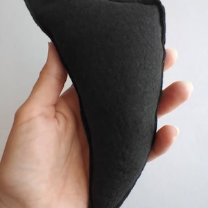 May include: A black, triangular-shaped felt object with a stitched border. The object is held in a hand against a white background. The felt appears to be a solid, dark color.
