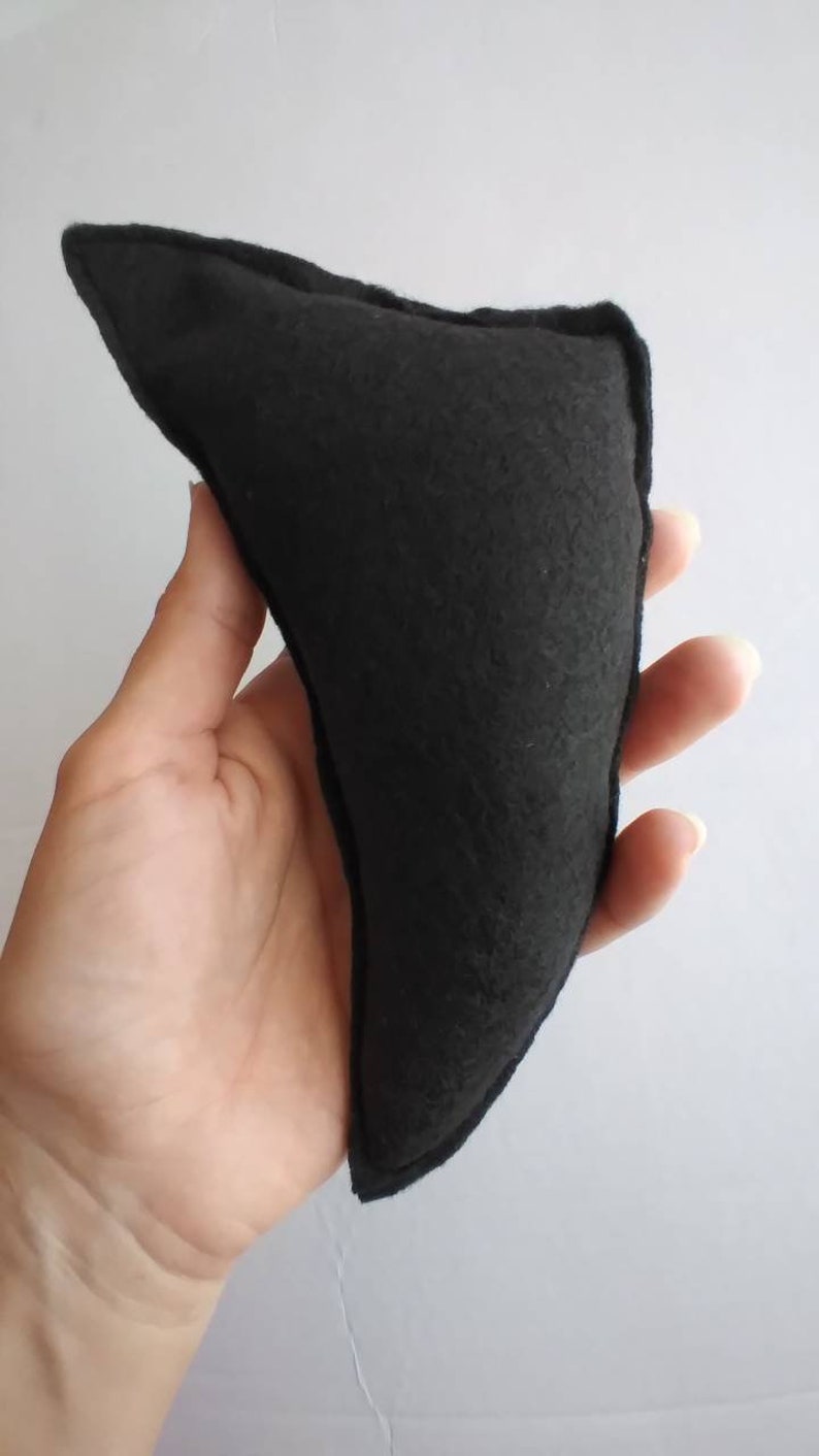 May include: A black, triangular-shaped felt object with a stitched border. The object is held in a hand against a white background. The felt appears to be a solid, dark color.