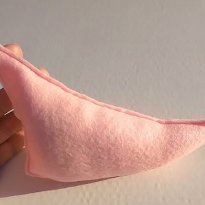 May include: A light pink, handmade felt elf ear. The ear is a soft, muted pink color and has a rounded shape with a pointed tip. The felt appears to be stitched together along the edges.