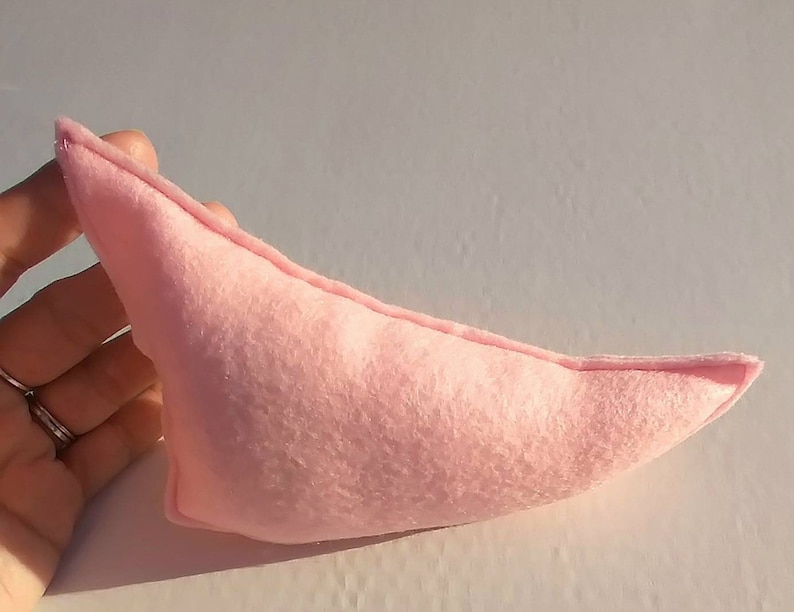 May include: A light pink, handmade felt elf ear. The ear is a soft, muted pink color and has a rounded shape with a pointed tip. The felt appears to be stitched together along the edges.
