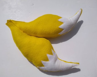 May include: Two plush yellow fox tails with white tips, lying on a white surface. The tails are made of soft fabric and are designed with a curved shape and a starburst pattern at the end.