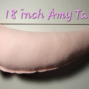 May include: A light pink, plush tail, approximately 18 inches long, with a curved shape and pointed tip. The text "18 inch Amy Tail" is written in a cursive font above the tail. The tail appears to be made of a soft fabric.