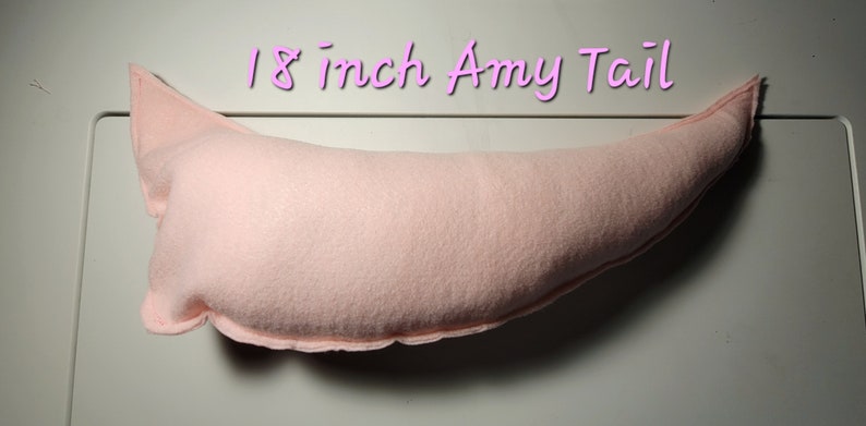 May include: A light pink, plush tail, approximately 18 inches long, with a curved shape and pointed tip. The text "18 inch Amy Tail" is written in a cursive font above the tail. The tail appears to be made of a soft fabric.
