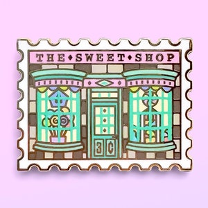 May include: Gold-colored enamel pin shaped like a postage stamp with the text "THE SWEET SHOP" and a cartoon image of a storefront with a pink awning and green windows.