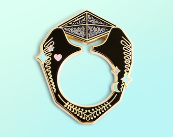 May include: Gold-tone enamel pin featuring a black diamond ring with a sparkly center, surrounded by stars and hearts. The ring is set against a light blue background.