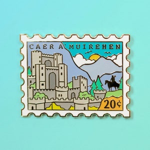 May include: A pin shaped like a postage stamp featuring a castle scene with the text "CAER A'MUIREHEN". The pin has a light blue sky, gray mountains, and a rider on horseback. The stamp reads "20¢".