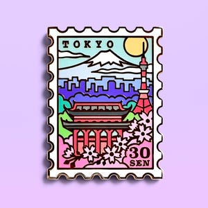 May include: A colorful enamel pin in the shape of a postage stamp, featuring a Japanese pagoda, Mount Fuji, and Tokyo Tower. The pin has the word "TOKYO" at the top and "30 SEN" at the bottom. The background is a light purple.