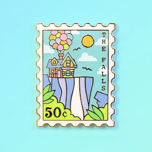 May include: A gold-colored enamel pin shaped like a postage stamp with a cartoon image of a house with balloons flying away from a cliff. The text "THE FALLS" is printed vertically on the right side of the stamp. The stamp also says "50¢".