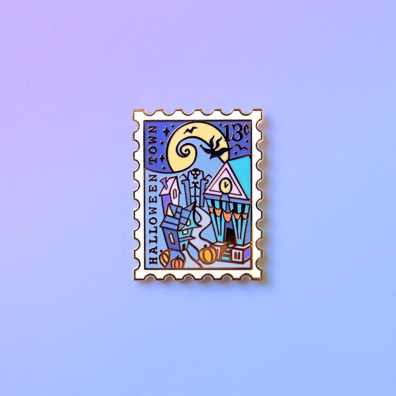 Op de afbeelding: Een witte emaille pin in de vorm van een postzegel met een zwarte omtrek. De pin heeft een cartoonillustratie van een stad met een klokkentoren, een scheve toren en een halve maan. De tekst "HALLOWEEN TOWN" staat verticaal aan de linkerkant van de pin. De tekst "13&cent;" staat aan de rechterkant van de pin.