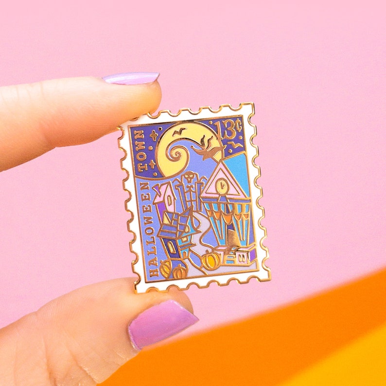 Op de afbeelding: Een witte emaille pin in de vorm van een postzegel met een gouden rand. De pin heeft een kleurrijke illustratie van de stad Halloween Town uit de film "The Nightmare Before Christmas". De tekst "HALLOWEEN TOWN" staat verticaal gedrukt aan de linkerkant van de pin. De tekst "13&cent;" staat gedrukt aan de rechterkant van de pin.