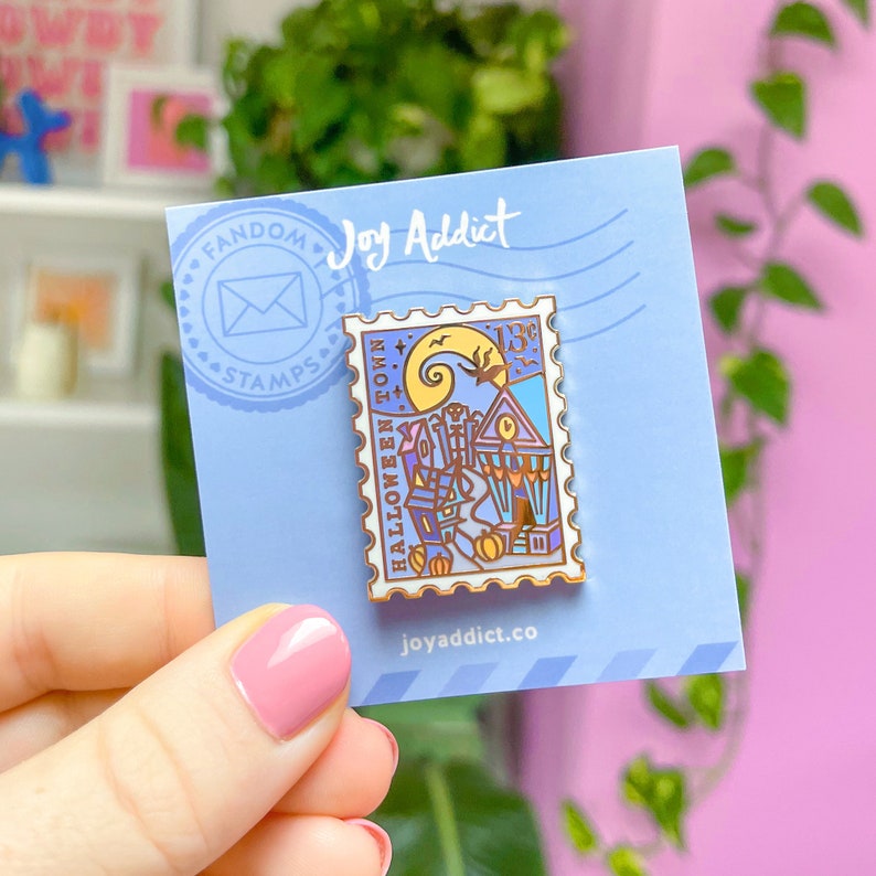 Op de afbeelding: Een blauwe en witte emaille pin in de vorm van een postzegel met de tekst "Halloween Town" en een 13&cent;-zegel. De pin heeft een cartoonafbeelding van een stad met een spookhuis en een jack-o-lantern.
