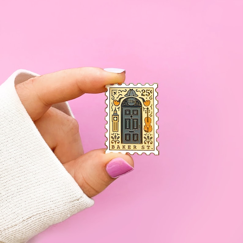 May include: A gold-colored enamel pin shaped like a postage stamp with a black door, a clock tower, a violin, and the text "25c BAKER ST."