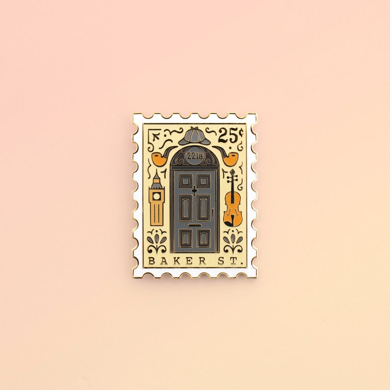 May include: A gold-colored enamel pin shaped like a postage stamp with a black and white illustration of a door with the number 221B above it. The pin also features a magnifying glass, a violin, and a clock tower. The text "BAKER ST." is printed below the door. The pin is on a light pink background.