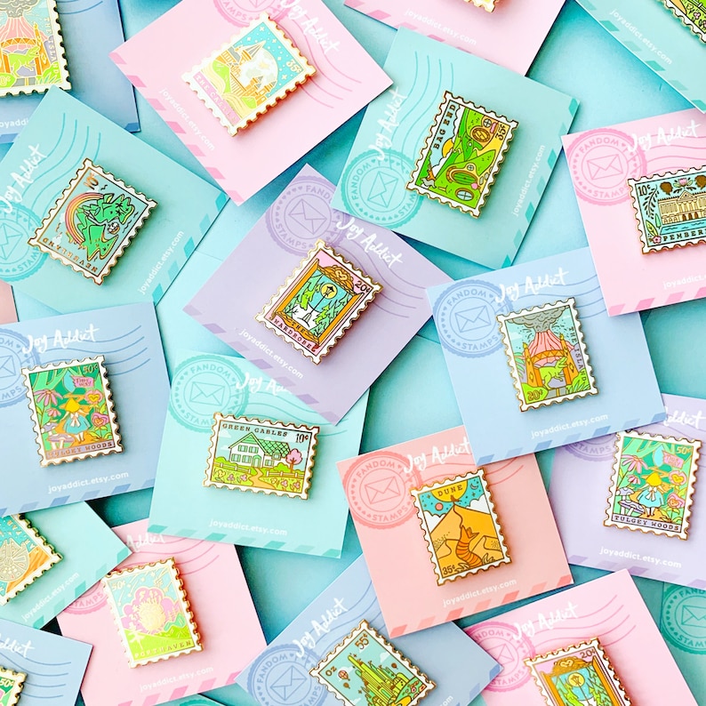 Op de afbeelding: Een verzameling kleurrijke emaille-pins in de vorm van postzegels. Elke pin heeft een andere illustratie, waaronder een groene kabelbaan, een vulkaan, een paddenstoelenbos en een duinlandschap. De pins worden getoond op pastelroze en blauwe kaarten met de tekst "Joy Addict" en "Fandom Stamps".