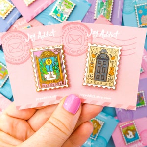 May include: Two gold-colored enamel pins with a vintage postage stamp design. The first pin features a pink and yellow design with the text "The Wardrobe" and the number "20". The second pin features a yellow and blue design with the text "Baker St." and the number "25".