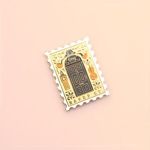 May include: A gold-colored enamel pin shaped like a postage stamp with a black door with the number "221B" on it. The pin also features a violin, a clock tower, and the words "BAKER ST."