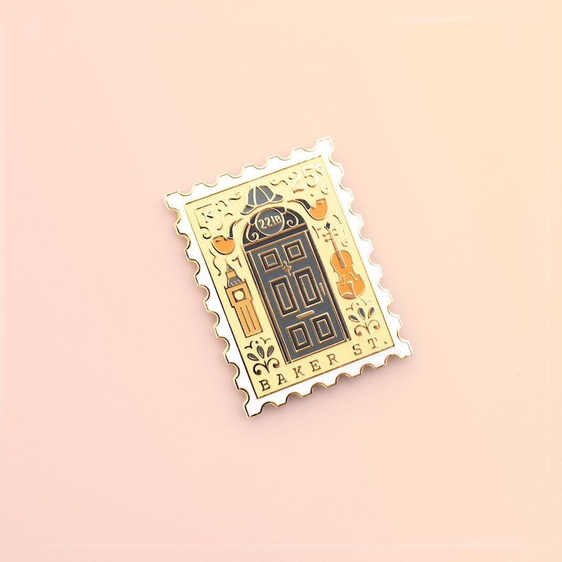May include: A gold-colored enamel pin shaped like a postage stamp with a black door with the number "221B" on it. The pin also features a violin, a clock tower, and the words "BAKER ST."