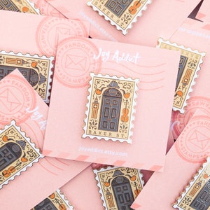 May include: A gold-colored enamel pin shaped like a postage stamp with a black door with gold accents and the text "221B BAKER ST."  The pin is surrounded by a white border with the text "FANDOM STAMPS" and "25".