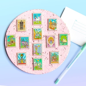 May include: A collection of 10 enamel pins featuring various whimsical illustrations, including a unicorn, a castle, a Totoro, and a cityscape. The pins are arranged on a pink corkboard and resemble postage stamps with different prices.