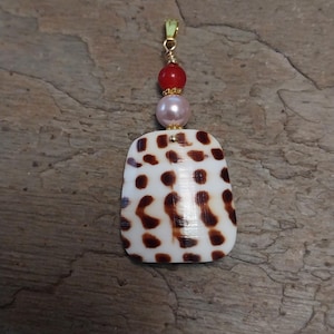 May include: A pendant featuring a white shell with brown spots, topped with a pink pearl and a red bead. The pendant is suspended from a gold-colored bail. The shell has a rectangular shape with rounded edges.