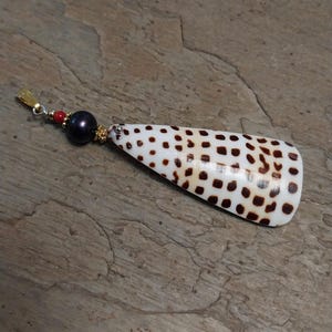 May include: A pendant featuring a white shell with brown spots, a dark pearl, and red and gold accents. The shell is elongated and has a unique pattern. The pendant is likely a piece of jewelry.