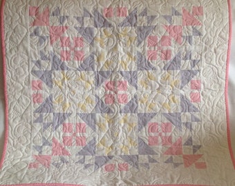 May include: A baby quilt with a patchwork design featuring pink, yellow, and grey squares and triangles. The quilt is quilted with a floral pattern.