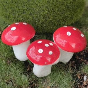 May include: Three decorative mushroom figurines with bright red caps and white polka dots. The mushroom stems are white. The mushrooms are arranged on a bed of green grass, with a large mossy green ball in the background.