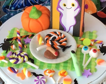 Könnte beinhalten: Eine Miniatur-Halloween-Dekoration auf einem weißen Tisch. Sie zeigt einen orangefarbenen Kürbis, eine Süßigkeitentüte mit einem Geist und verschiedene Süßigkeiten wie Lutscher und Zuckermais. Schwarze Spinnen und Fledermäuse sind auf dem Tisch verstreut.