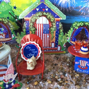 May include: A miniature patriotic scene with a red metal chair, a small house with an American flag door, and a sign that reads "One Nation Under God." The scene includes a small table with drinks and a dessert, and a sign that says "4th of July."